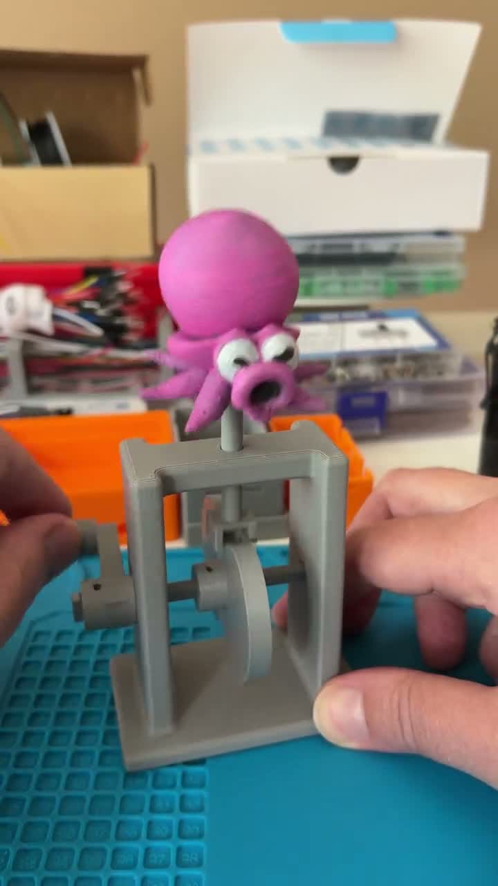 A hand slowly cranks a 3D-printed automata: a magenta Octorok (the Zelda octopus) bobs up and down on a grey printed frame.