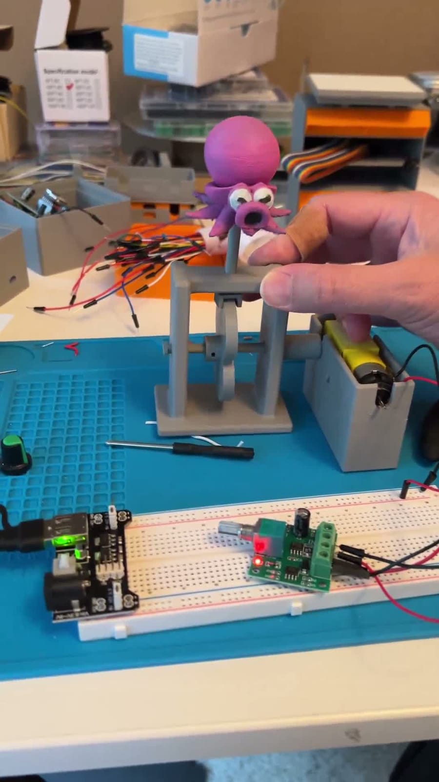 A 3D-printed automata on a workbench, a magenta Octorok bobbing on a grey frame, driven by a small motor wired to a breadboard and battery pack beside it.