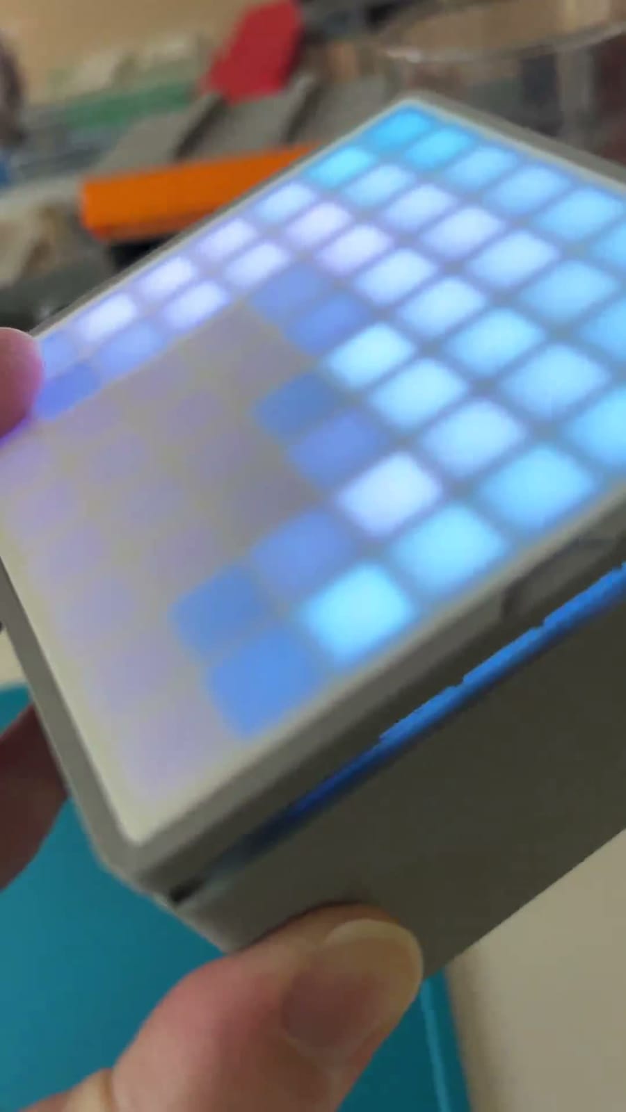 A hand holding a small square LED tile in a white printed enclosure, its 8x8 grid glowing in soft blue and lavender through a diffuser, with no cable attached.