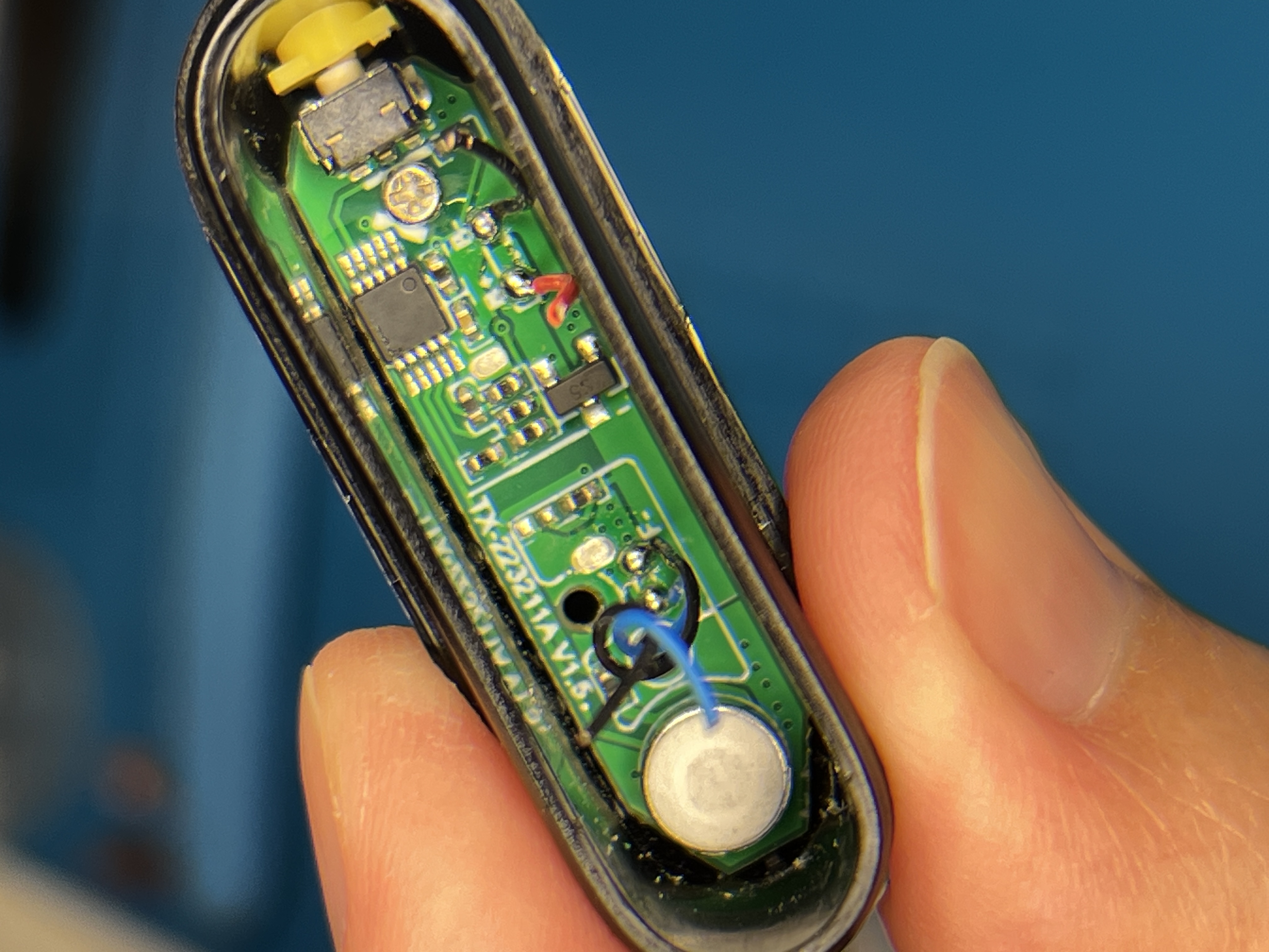 A small black vape shell held in fingers and cracked open, showing the green circuit board inside: a silver USB-C connector and a cross-head screw at the top, a black square chip in the middle, and a white disc wired with two fine leads at the bottom.