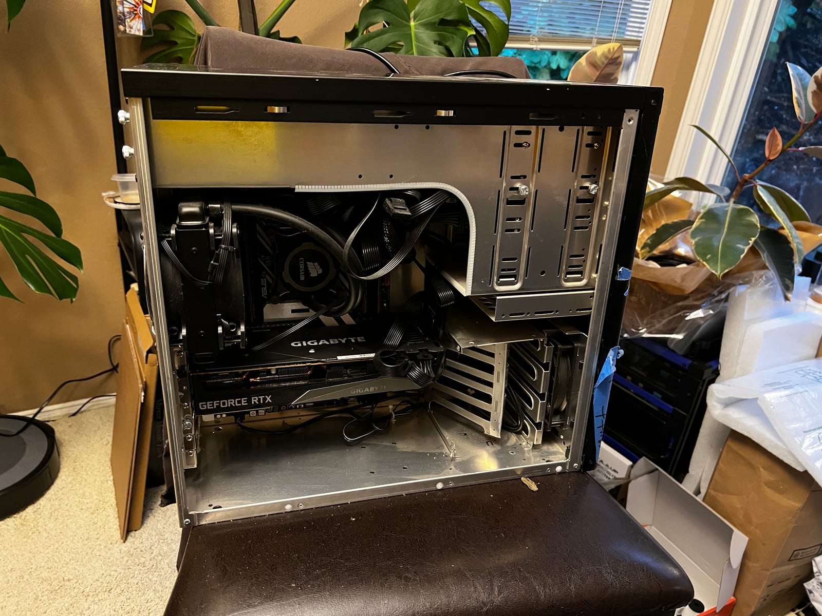 A black computer tower with its side panel off, lying open on a dark leather chair in a room full of houseplants. Inside, a Corsair liquid cooler and a large Gigabyte GeForce RTX graphics card sit above an empty lower bay, with a braided cable routed around the drive cage.